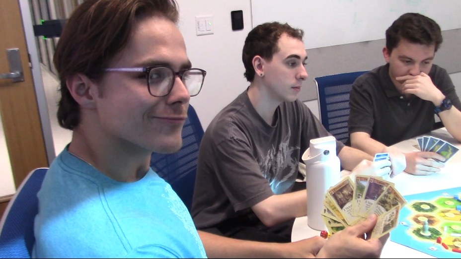 Max, Cody, and Brian sitting at a table playing a board game; Max has good cards at his hand and looks at the camera smugly. 